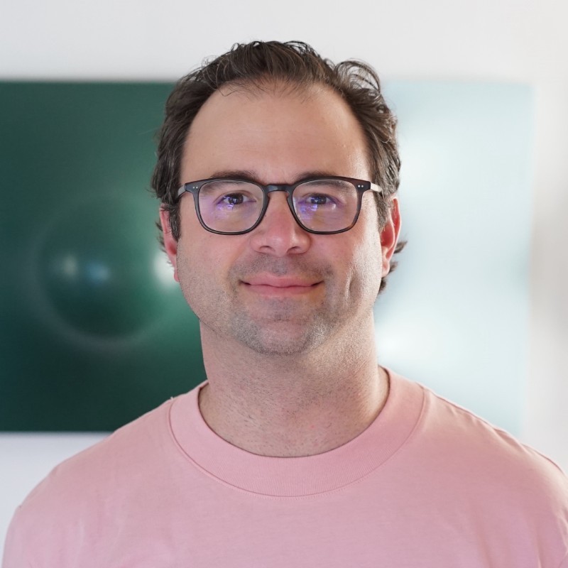 Portrait of a smiling man wearing glasses and a pink shirt, standing in front of a green backdrop.