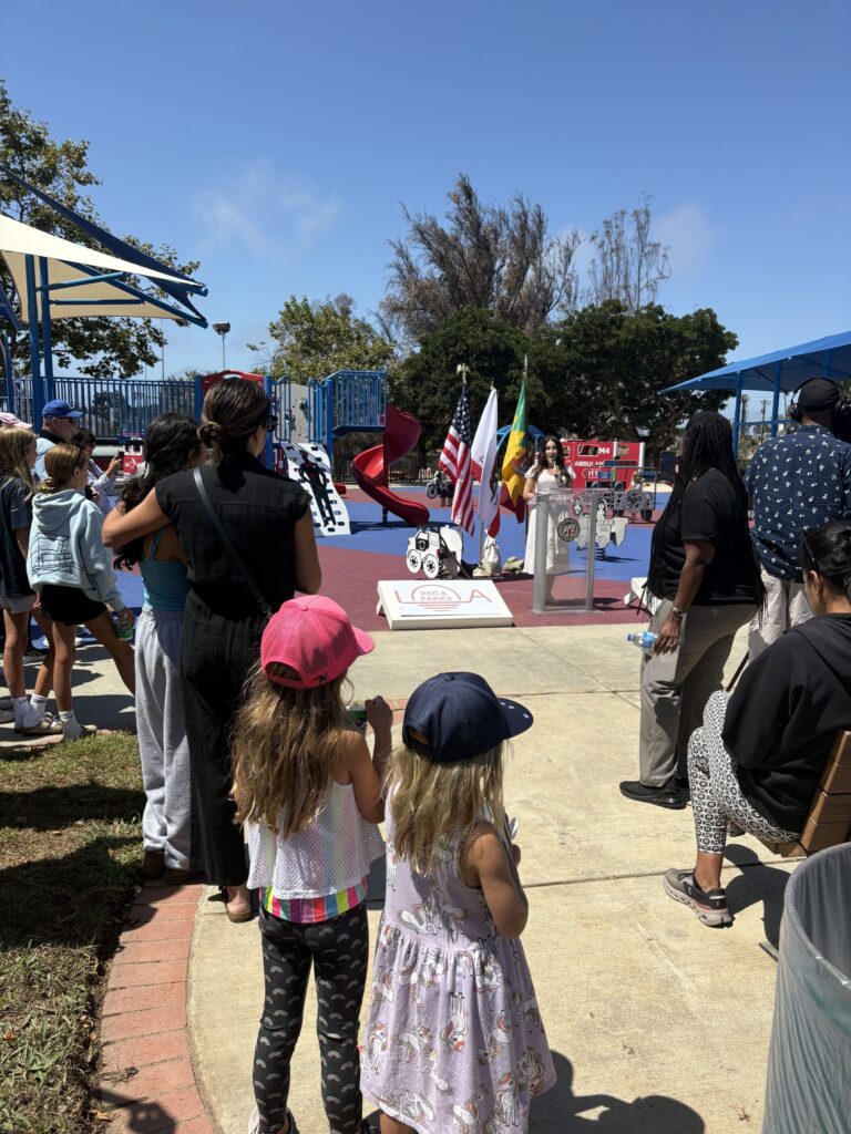 A group of children and adults at a playground event, with a speaker at a podium surrounded by flags. Play equipment and trees are visible in the background.